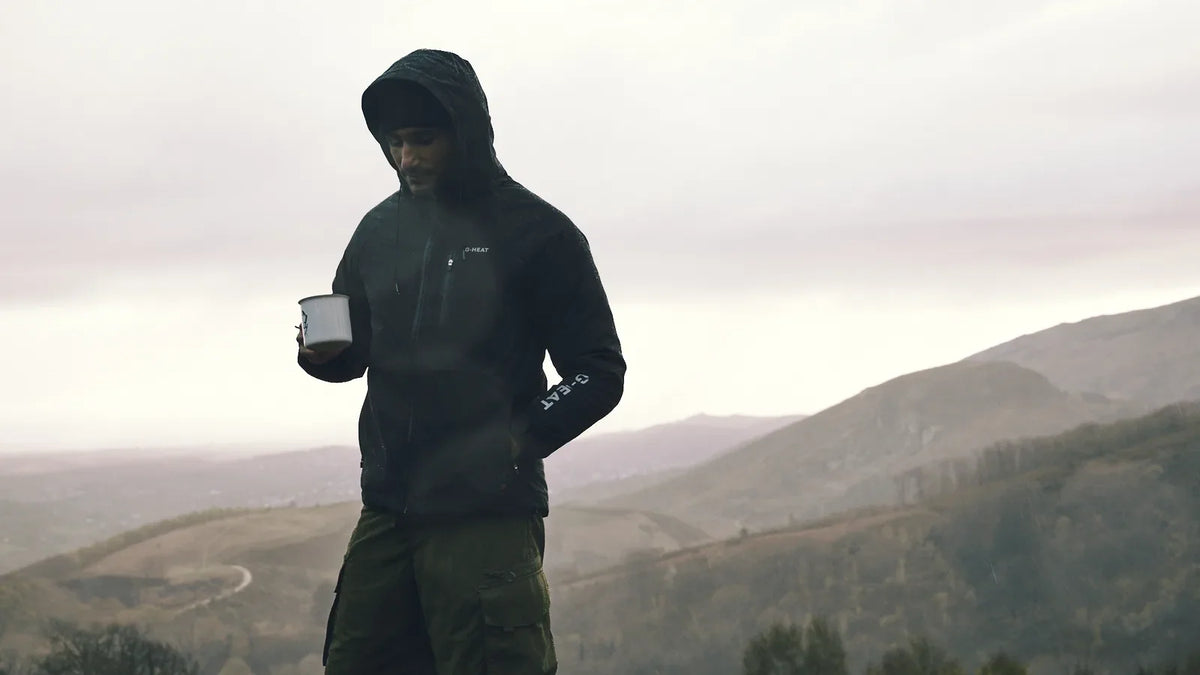 randonneur en veste à capuche tenant une tasse face aux collines embrumées
